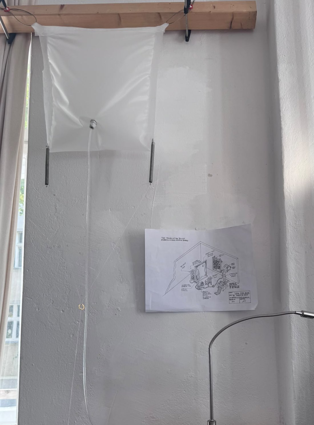 Prototype membrane suspended from a wooden beam on springs, with an air inlet at its centre; the isometric drawing hangs on the wall beside it